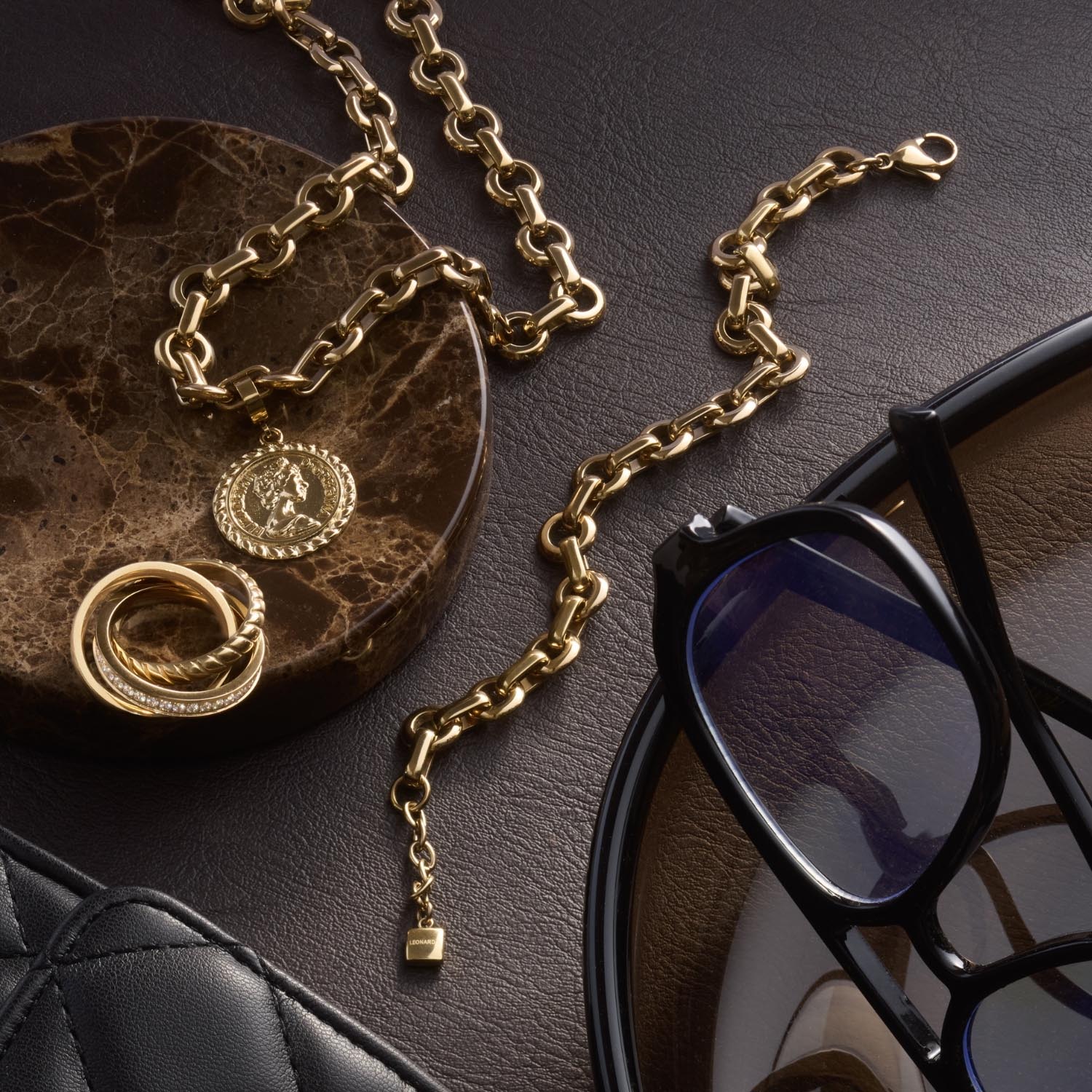 Goldenes Armband NALA mit Kette, Ringen und Anhängern auf Dekotablett neben Sonnenbrille