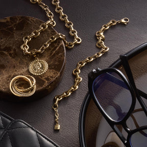 Goldenes Armband NALA mit Kette, Ringen und Anhängern auf Dekotablett neben Sonnenbrille