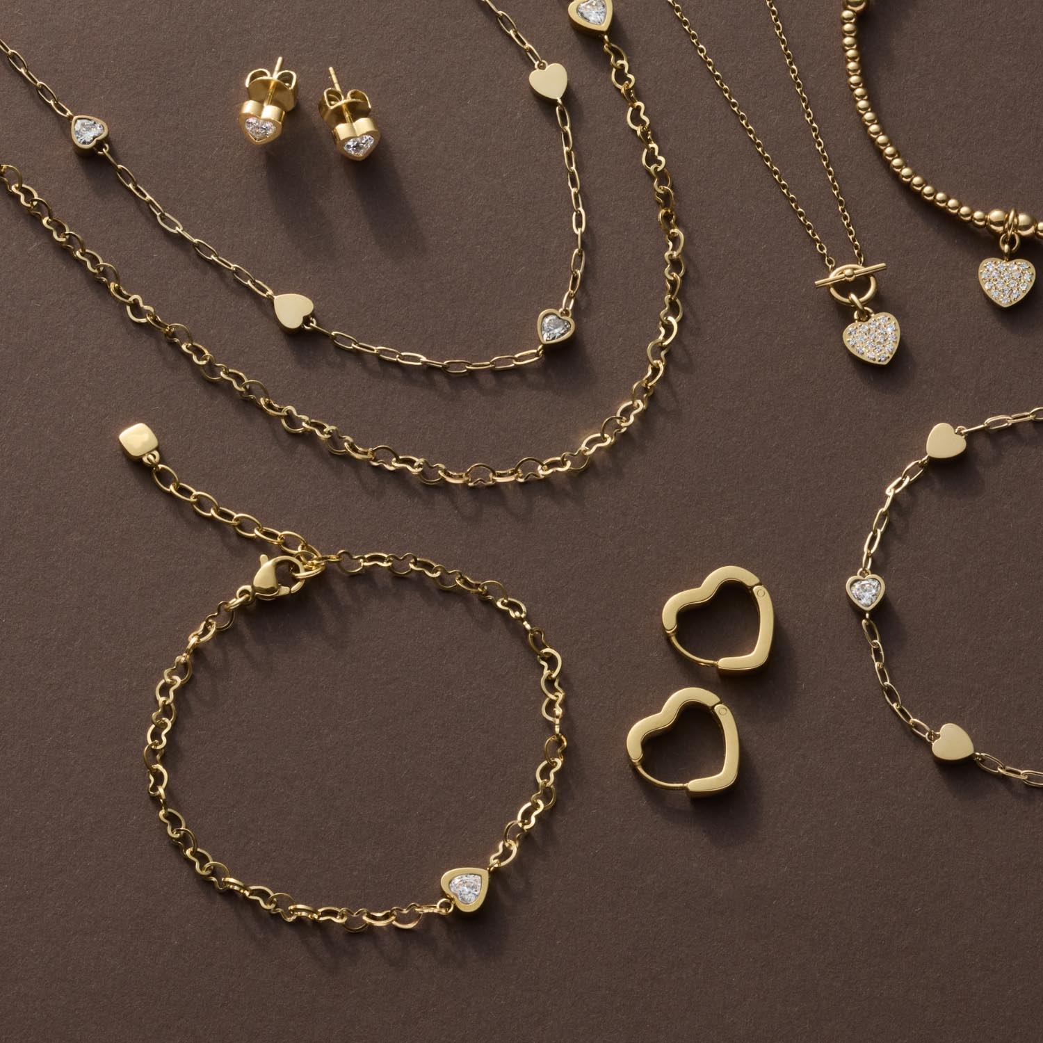Goldene Schmuckkollektion mit Halsketten, Armband JULY CIAO, Ohrringen und Herzanhängern auf braunem Hintergrund