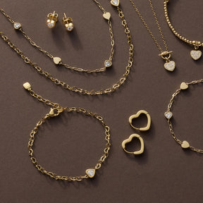 Goldene Schmuckkollektion mit Halsketten, Armband JULY CIAO, Ohrringen und Herzanhängern auf braunem Hintergrund