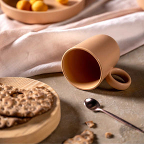 Keramiktasse ROMA apricot auf Leinentuch mit Keksen und Löffel, Obstschale im Hintergrund