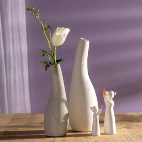 Weiße Keramikfiguren der Serie LUMINOSA auf Holzoberfläche vor violettem Hintergrund, daneben Vasen mit weißer Tulpe