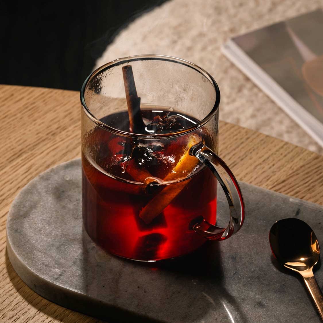 Teeglas NOVO mit dunklem Tee und Eiswürfeln auf grauem Tablett, Löffel daneben