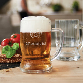 Bierseidel TAVERNA mit Bier gefüllt auf Holztisch, Gravur sichtbar, Tomaten und Brezel daneben