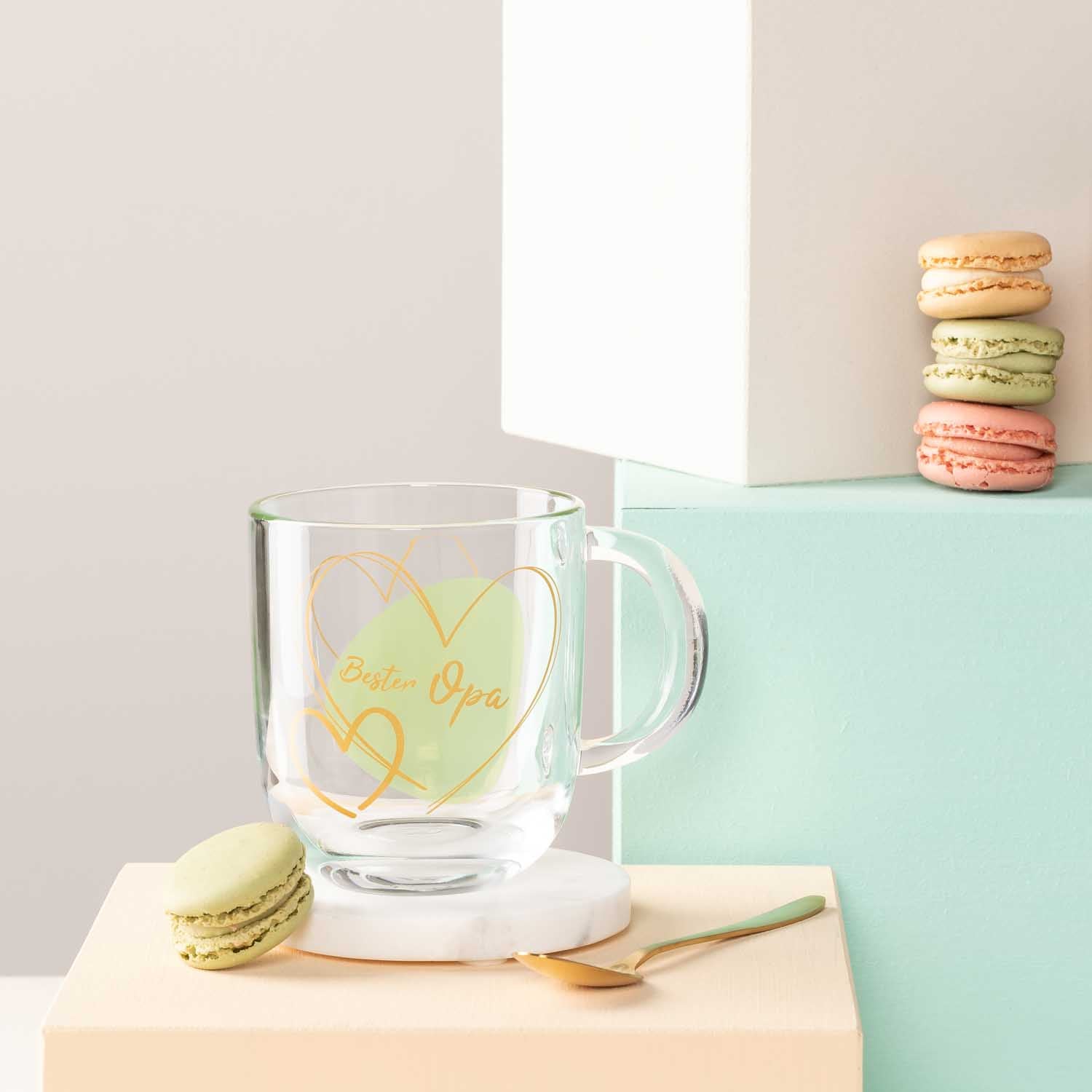 Glastasse EMOZIONE mit grüner Gravur "Bester Opa", daneben Makronen und Holzlöffel auf farbiger Ablage