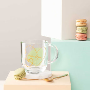Glastasse EMOZIONE mit grüner Gravur "Bester Opa", daneben Makronen und Holzlöffel auf farbiger Ablage