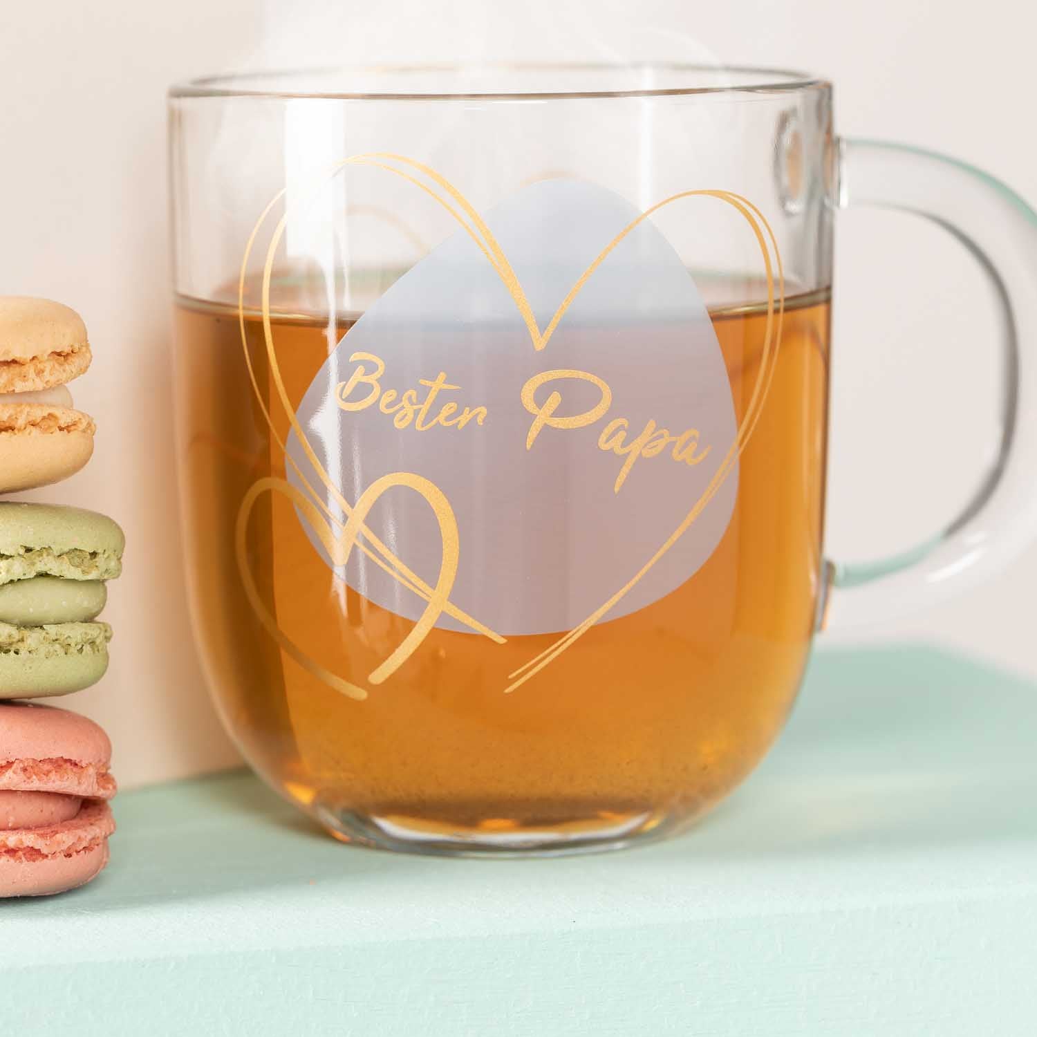 Glastasse mit Gravur "Besten Papa", gefüllt mit braunem Tee, neben bunten Macarons