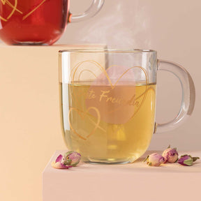 Glastasse mit Gravur gefüllt mit gelbem Tee, daneben getrocknete Rosenblüten