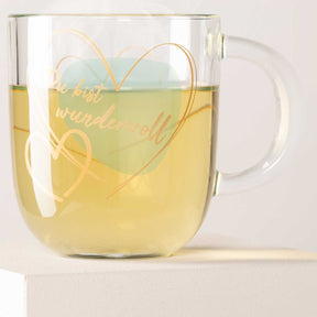 Glastasse EMOZIONE gefüllt mit gelblicher Flüssigkeit, Gravur mit Herz und Text auf der Oberfläche