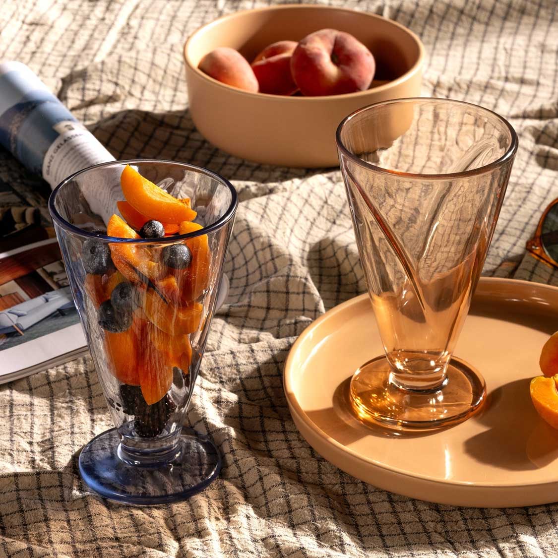 Eisbecher CONO gefüllt mit Getränk und Früchten auf sonnigem Terrassentisch, daneben Obstschale und leeres Glas