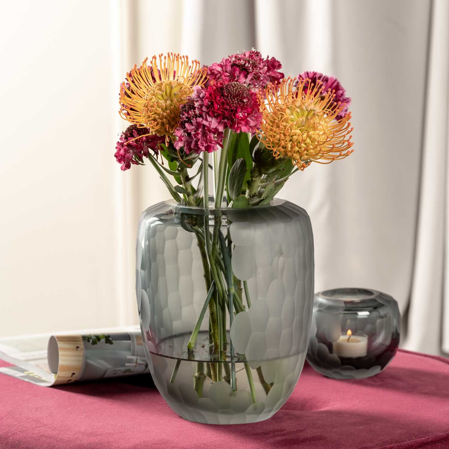 Vase BELLAGIO mit bunten Blüten auf rosa Tuch, Kerzen und Deko daneben