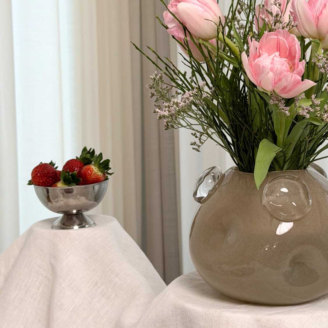 Vase HAZEL mit rosa Blüten und Gräsern, daneben Schale mit Erdbeeren auf Tuch