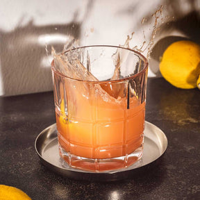 Whiskyglas mit spritzender orangefarbener Flüssigkeit, daneben Zitronen auf dunkler Oberfläche