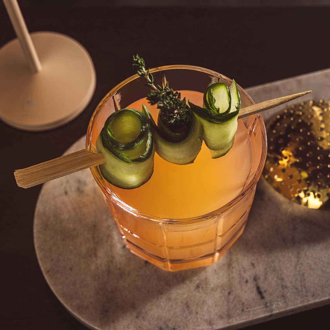 Whiskyglas gefüllt mit orangefarbenem Cocktail, garniert mit Gurkenscheiben und Thymian auf Holzstab