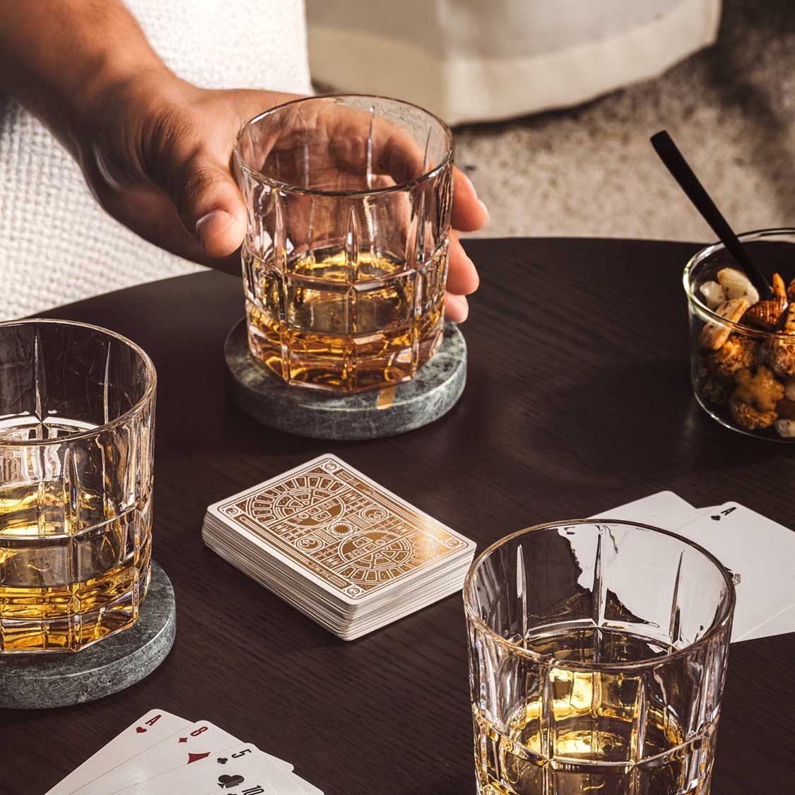 Whiskygläser mit Whiskey gefüllt auf schwarzem Tisch, Hand hält ein Glas, Spielkarten daneben