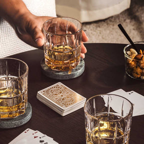 Whiskygläser mit Whiskey gefüllt auf schwarzem Tisch, Hand hält ein Glas, Spielkarten daneben