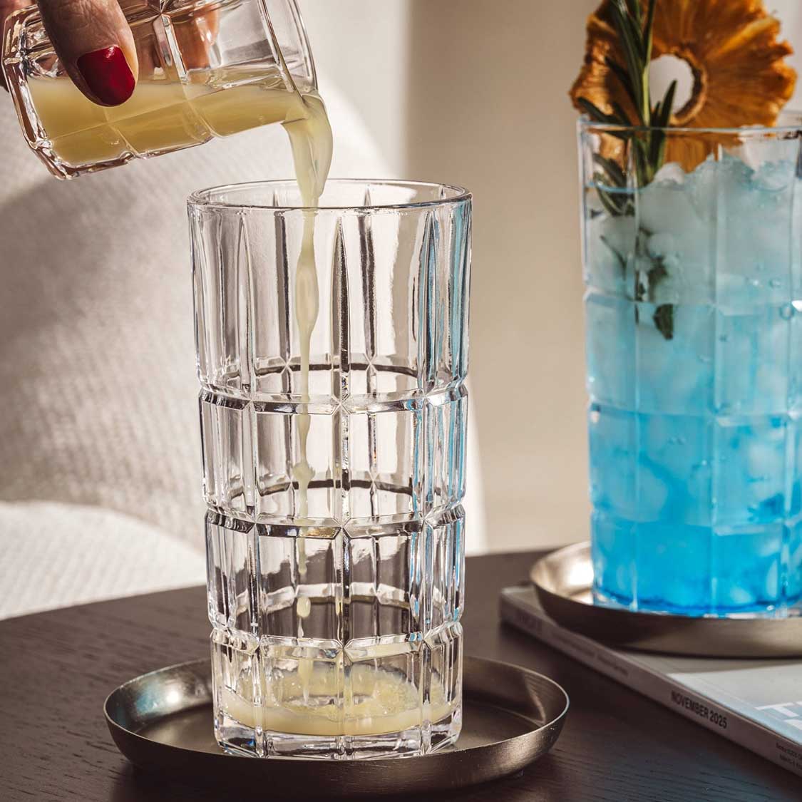 Longdrinkglas SPIRITII NOVA wird mit Getränk gefüllt, daneben Vase mit Blume und blaues Getränk