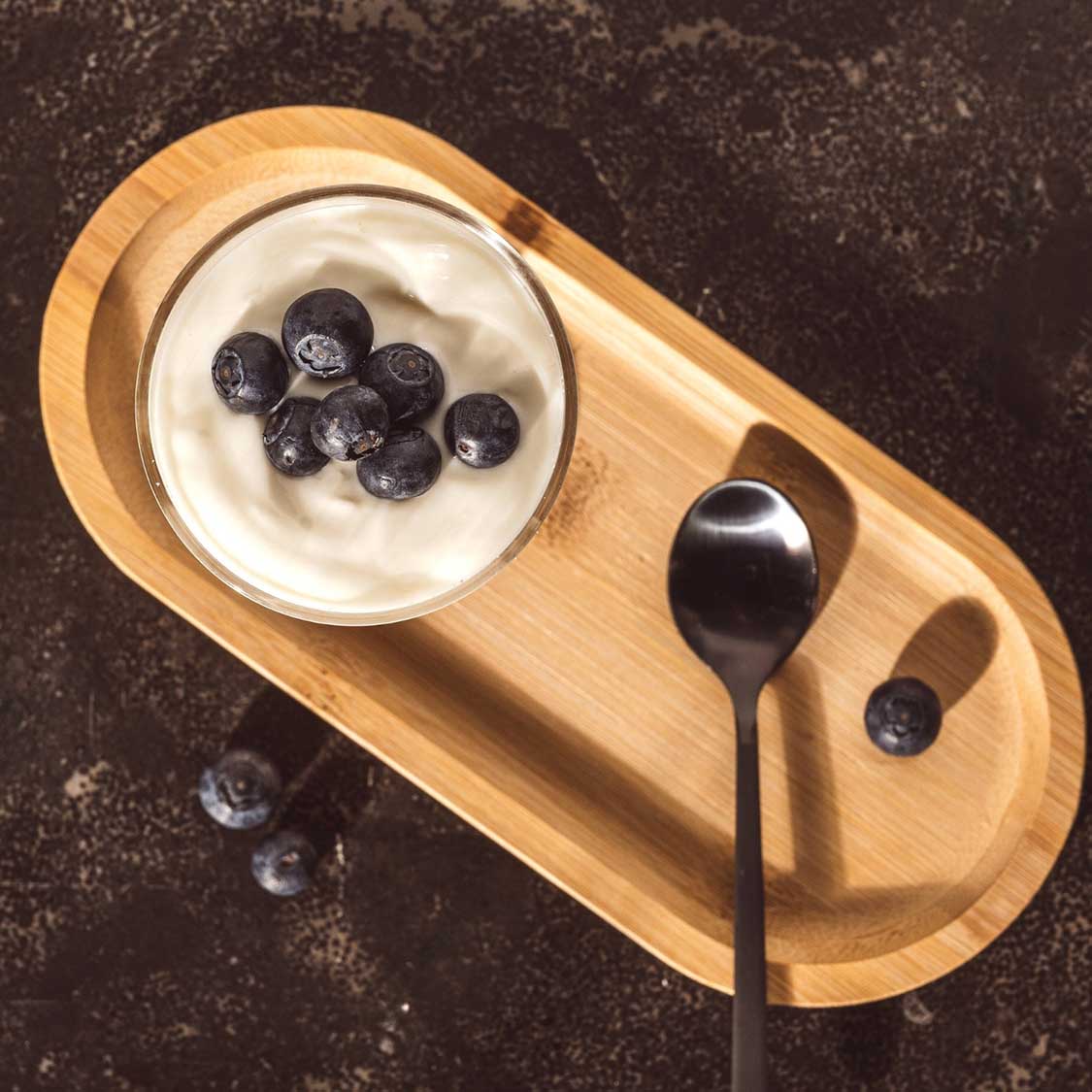 Holzbrett mit Joghurt, Blaubeeren und schwarzem Löffel, Stillleben
