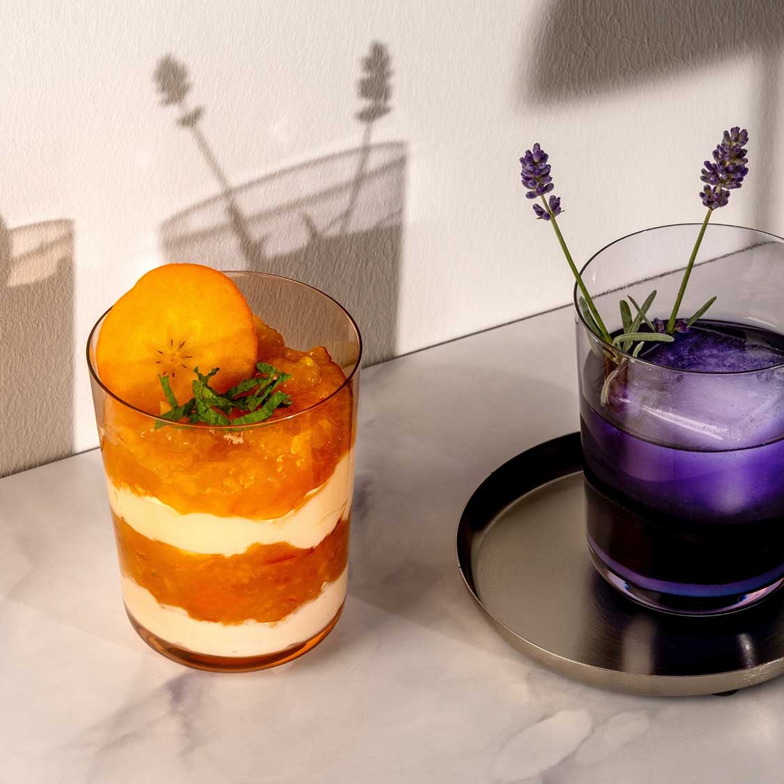 Trinkglas LEGGERO mit geschichtetem Cocktail und Orangenscheibe, neben violettem Getränk mit Lavendel