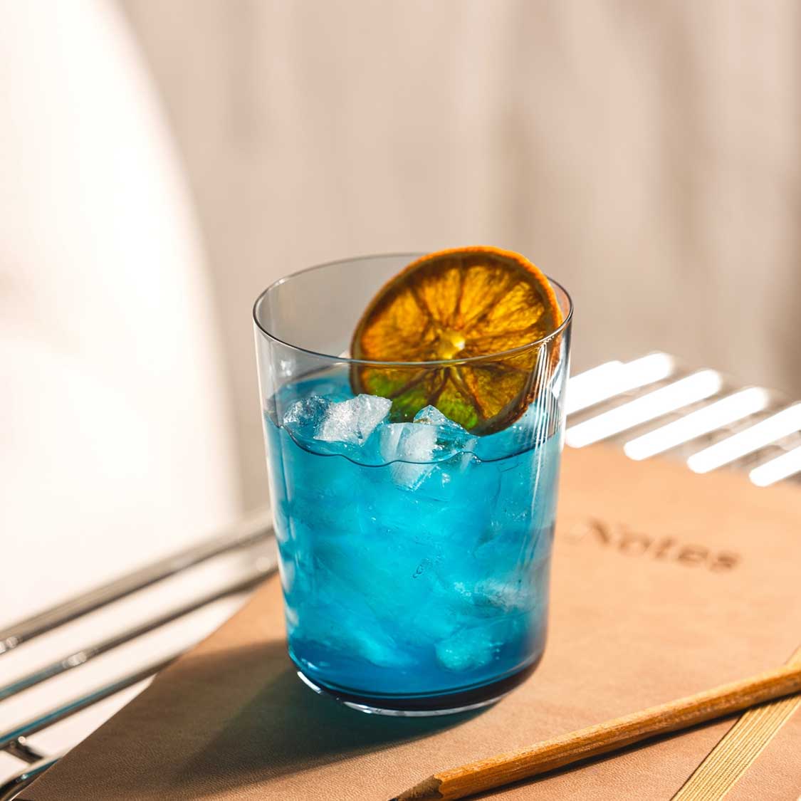 Trinkglas LEGGERO mit blauem Cocktail, Eiswürfeln und getrockneter Orangenscheibe auf Holztisch