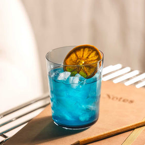 Trinkglas LEGGERO mit blauem Cocktail, Eiswürfeln und getrockneter Orangenscheibe auf Holztisch