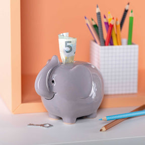 Spardose BAMBINI grau Elefant mit Geldschein, umgeben von Stiften und Bleistiften