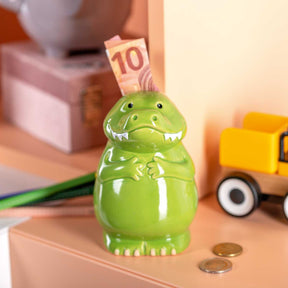 Spardose BAMBINI Krokodil grün mit Geldschein, Münzen und Spielzeugen auf Schreibtisch