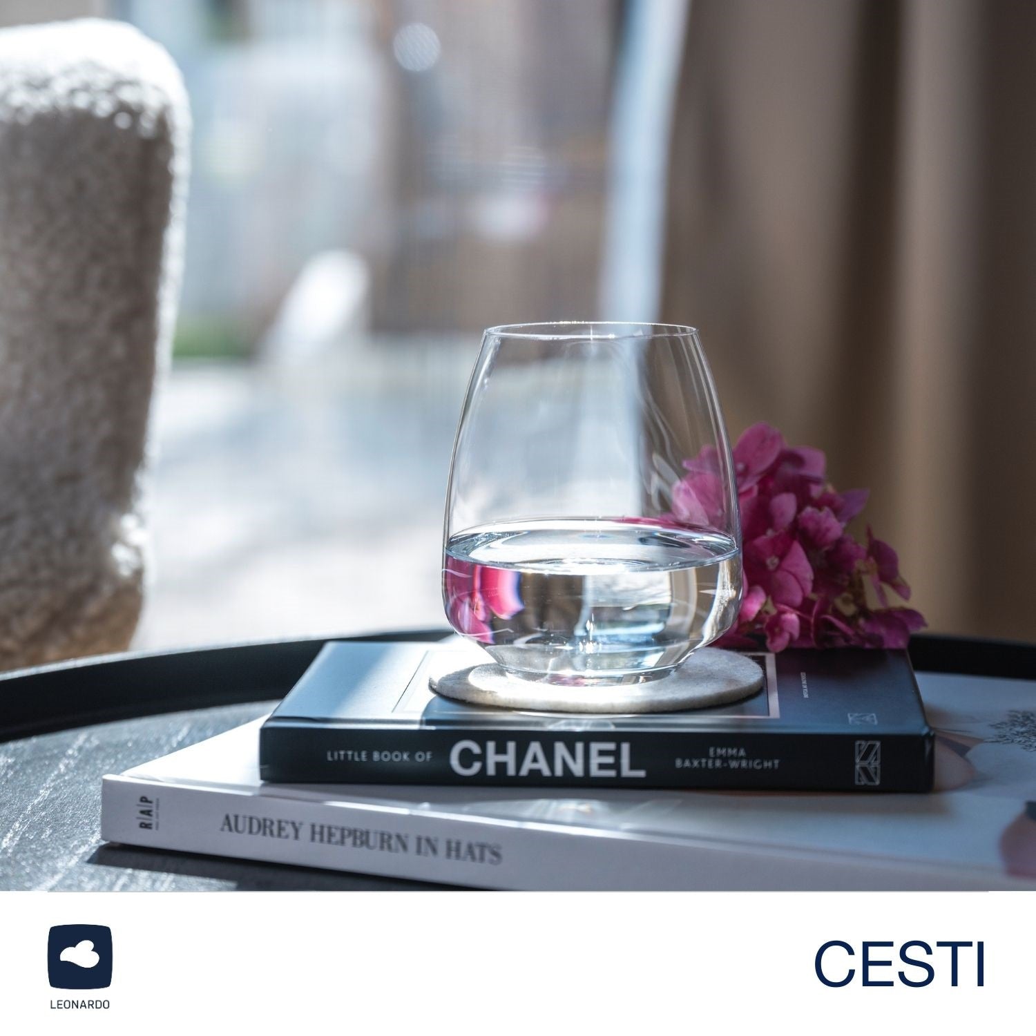 Trinkglas CESTI mit Wasser und Blüte auf Stapel mit Chanel-Buch, Wohnzimmer