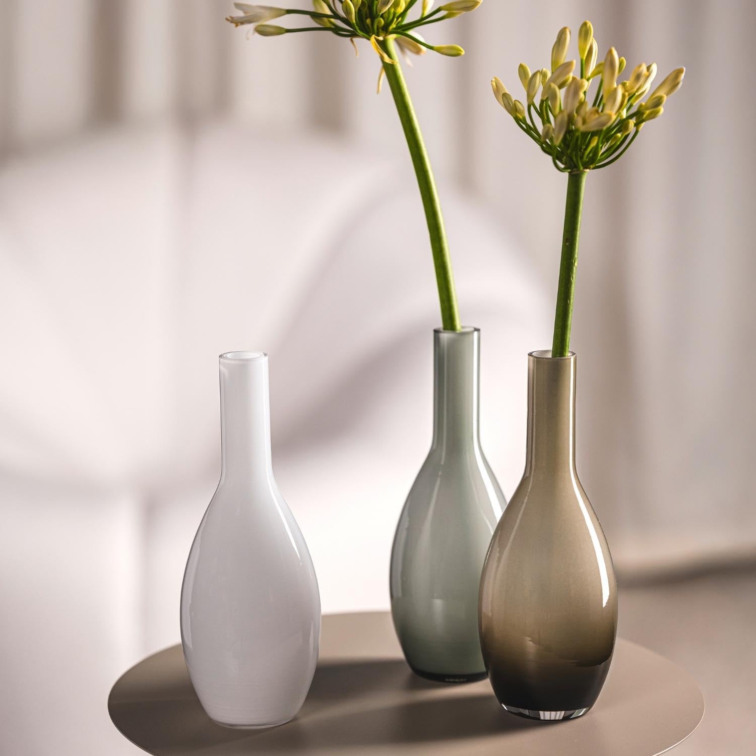 Drei Vasen mit Blüten in weißem, grauem und braunem Glas auf Tisch