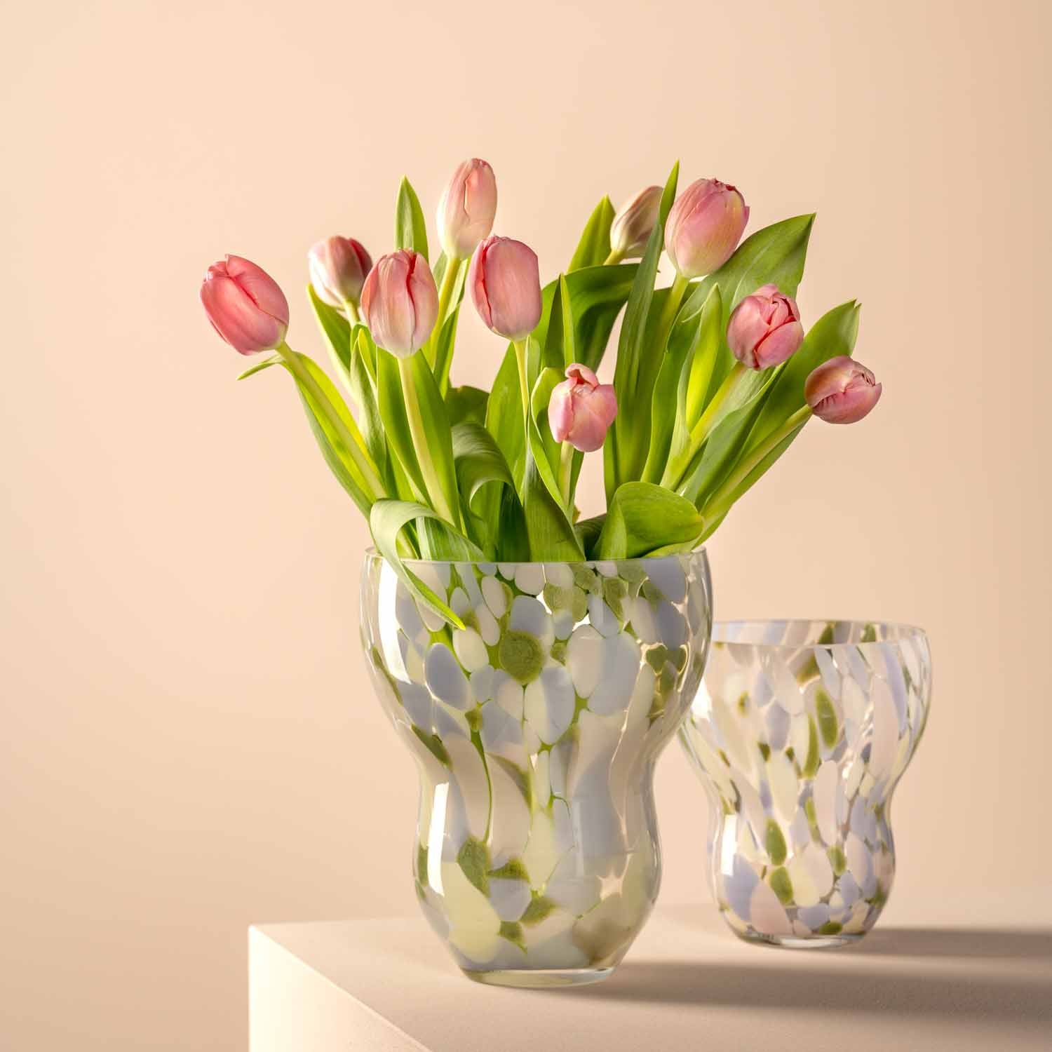 Vase VIVACE mit rosa Tulpen und grünen Blättern, daneben kleinere Vase, auf beigem Hintergrund