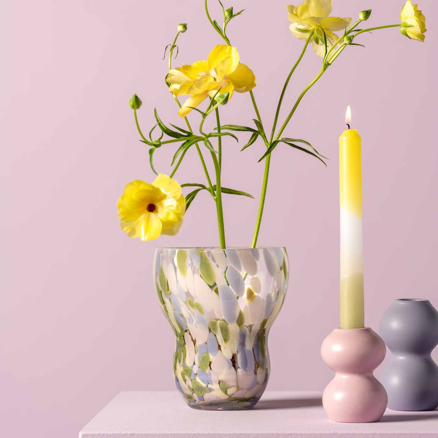 Vase VIVACE mit gelben Blüten, Kerzen und rosa Kerzenständer auf Tisch vor rosa Hintergrund