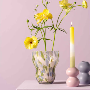 Vase VIVACE mit gelben Blüten, Kerzen und rosa Kerzenständer auf Tisch vor rosa Hintergrund
