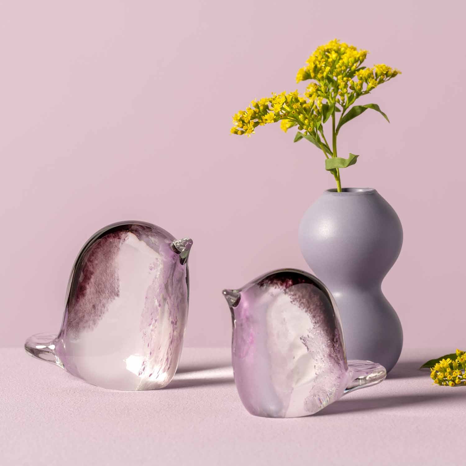 Zwei rosé Glasvögel und eine graue Vase mit gelben Blüten auf rosa Hintergrund