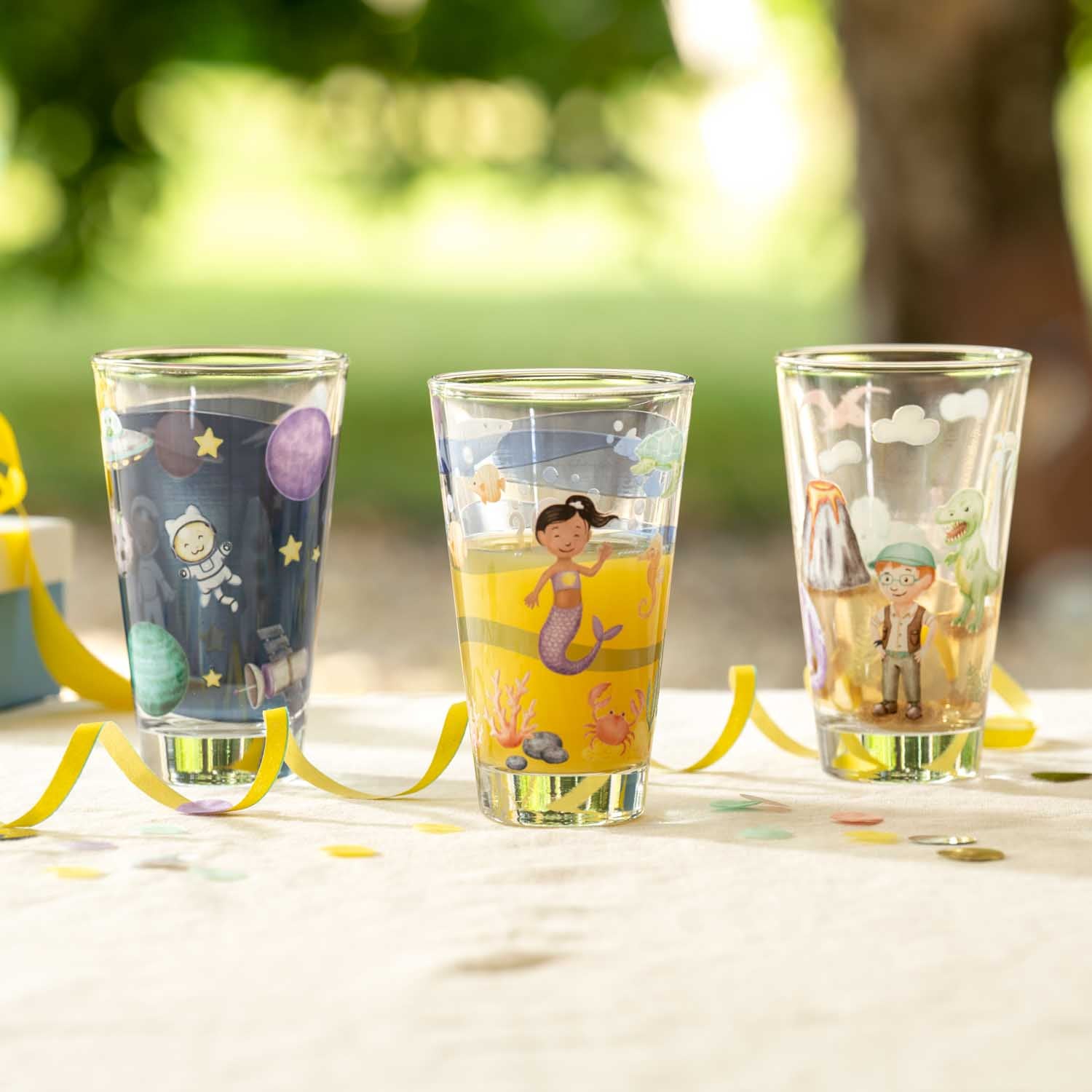 Drei bedruckte Trinkgläser BAMBINI AVVENTURA mit Kindermotiven auf Gartentisch im Freien