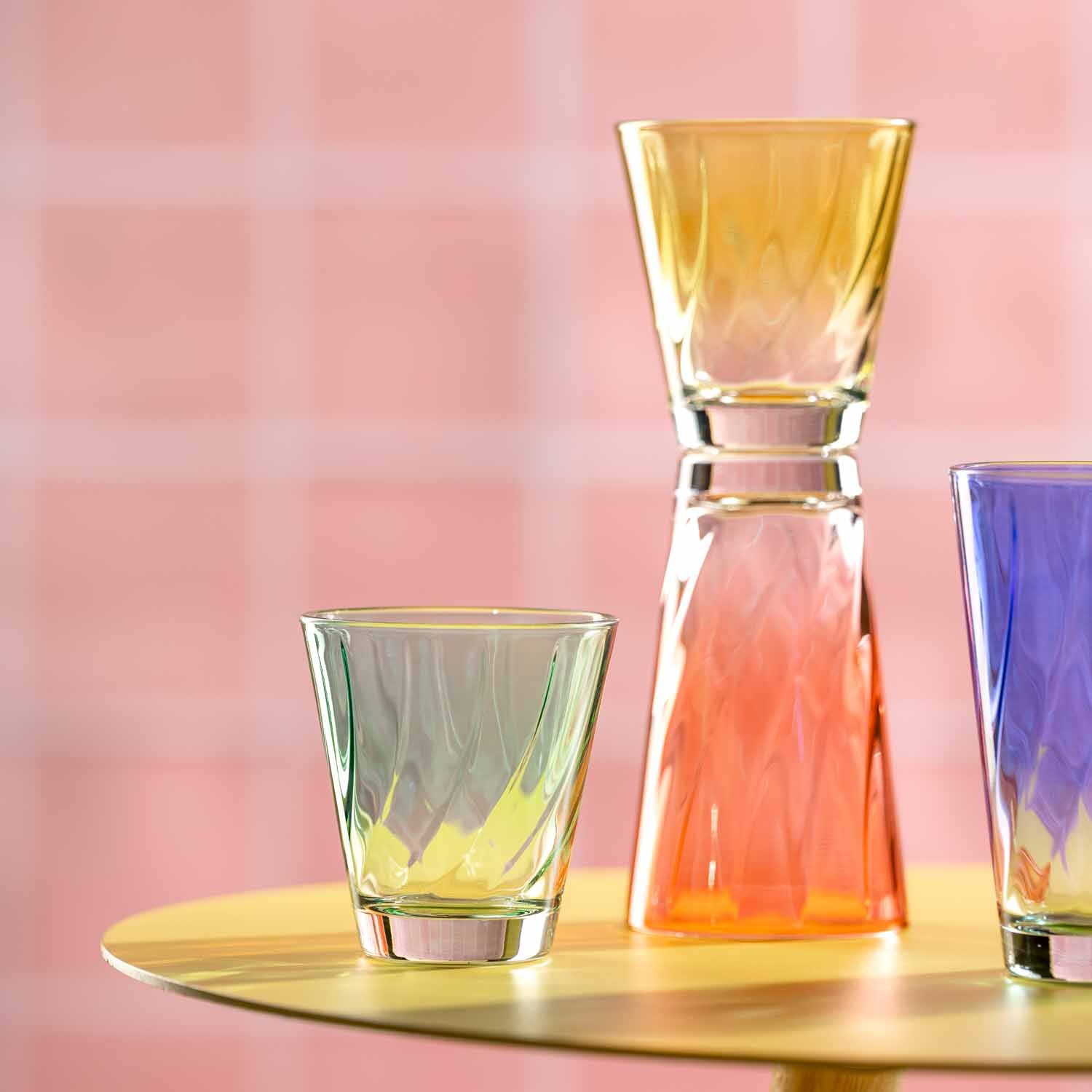 Trinkgläser TWIST in Gelb, Orange und Violett auf goldener Oberfläche vor rosa Hintergrund