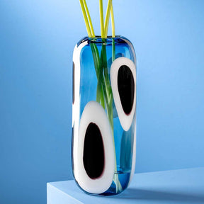 Vase OCCHI BLU mit weißen Augen und grünen Halmen vor blauem Hintergrund