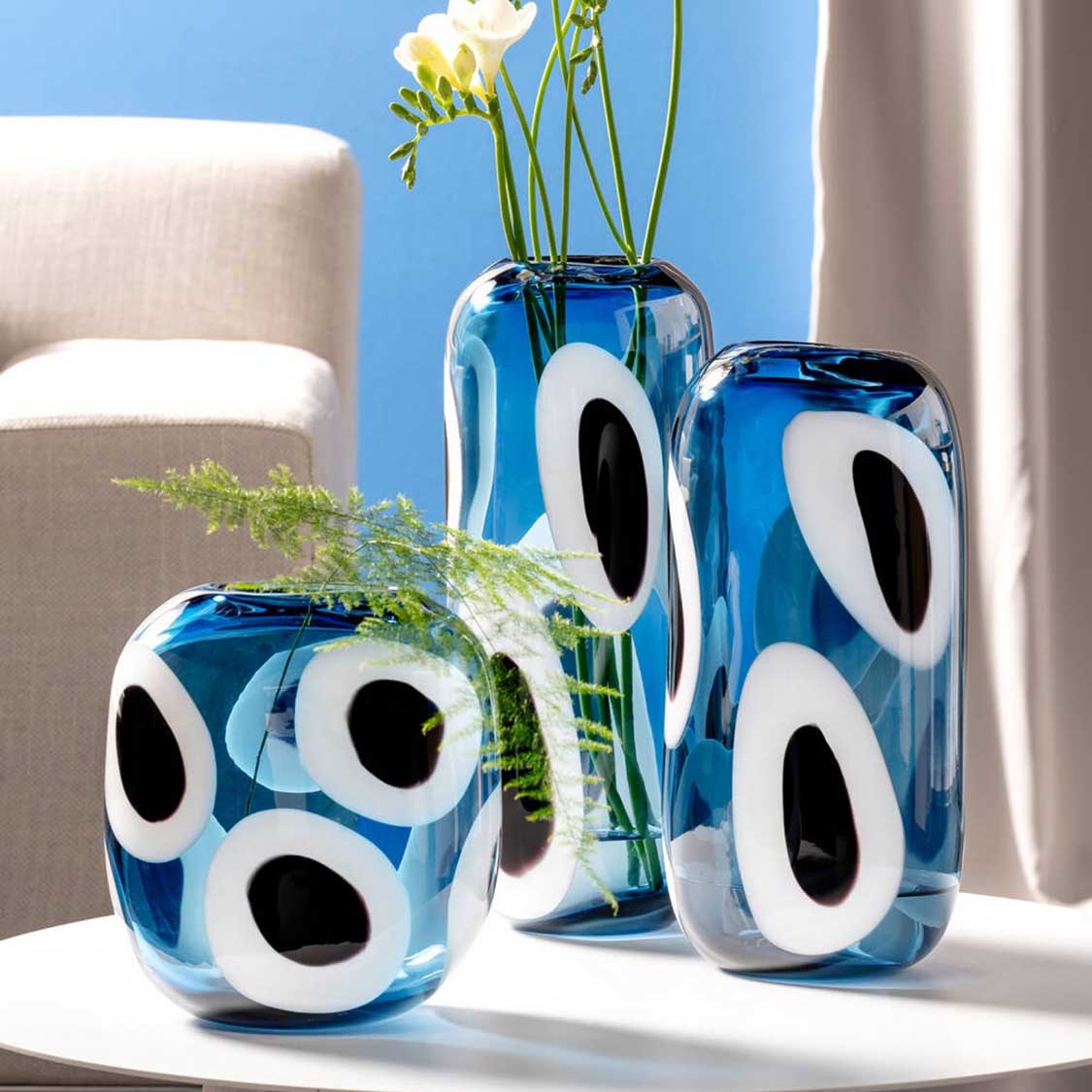 Blaue Glasvasen OCCHI BLU mit weißen und schwarzen Augen-Motiven, mit Blumen und Grünpflanzen dekoriert