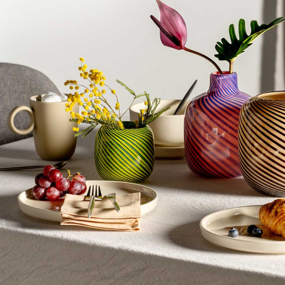 Grüne Vase FILO COLORI mit gelben Blüten auf gedecktem Tisch zwischen anderen Vasen und Tellern