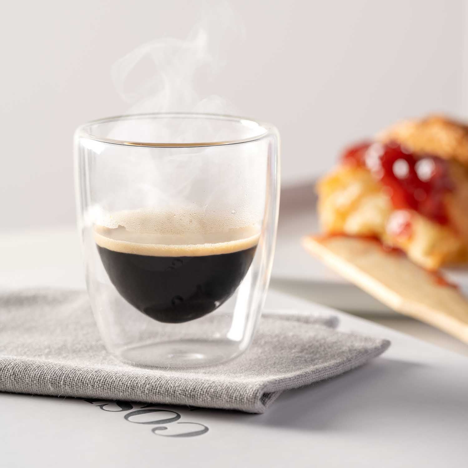 Doppelwandbecher Espresso DUO mit Espresso auf Leinentuch, Gebäck im Hintergrund