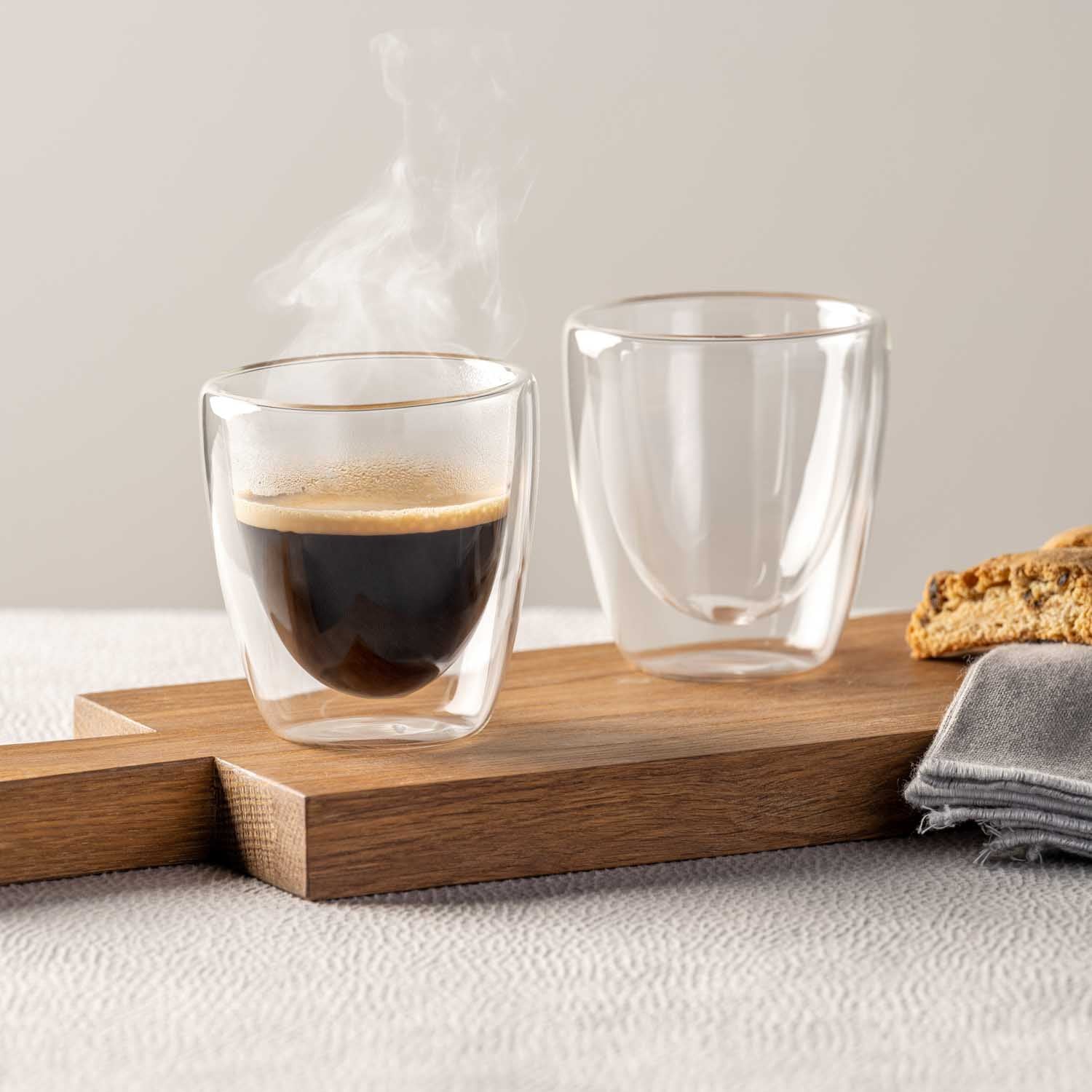 Doppelwandbecher Espresso DUO mit heißem Kaffee und leerem Becher auf Holzbrett, daneben Gebäck und Tuch