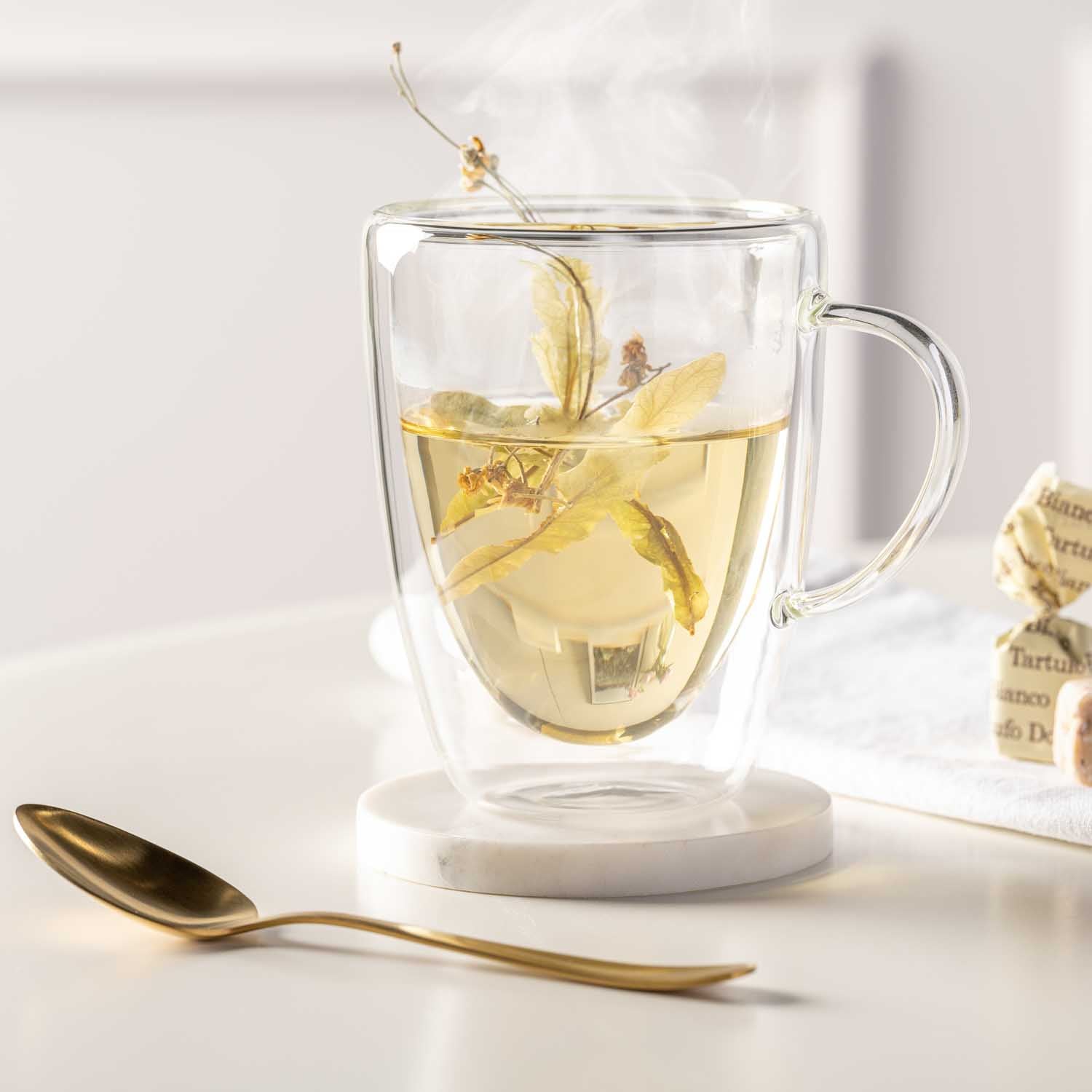 Doppelwandtasse DUO mit Kräutertee, goldener Löffel und Untersetzer auf Schreibtisch