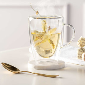 Doppelwandtasse DUO mit Kräutertee, goldener Löffel und Untersetzer auf Schreibtisch