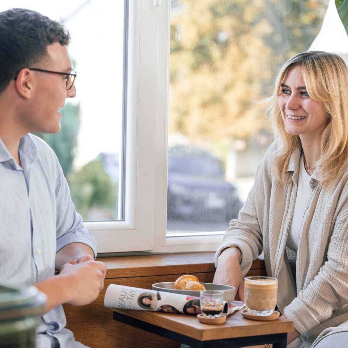 Zwei Personen an Fenster mit Espressogläsern GOCCE und Gebäck auf Holztisch
