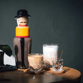 Espressoglas GOCCE mit Wabenmuster neben anderen Gläsern auf Holztisch, Dekofigur im Hintergrund