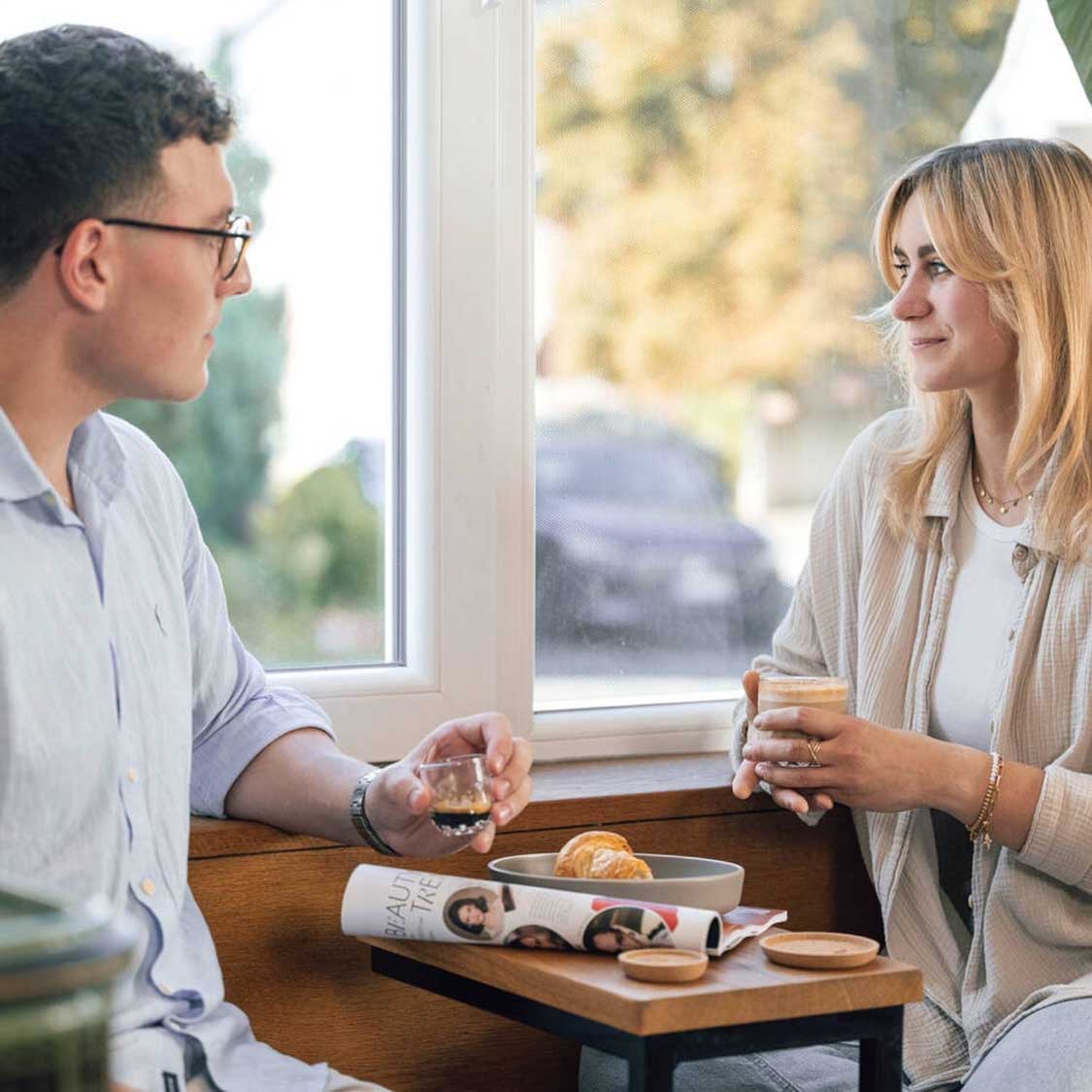 Zwei Personen an Fenster mit Espressogläsern und Gebäck auf Holztisch