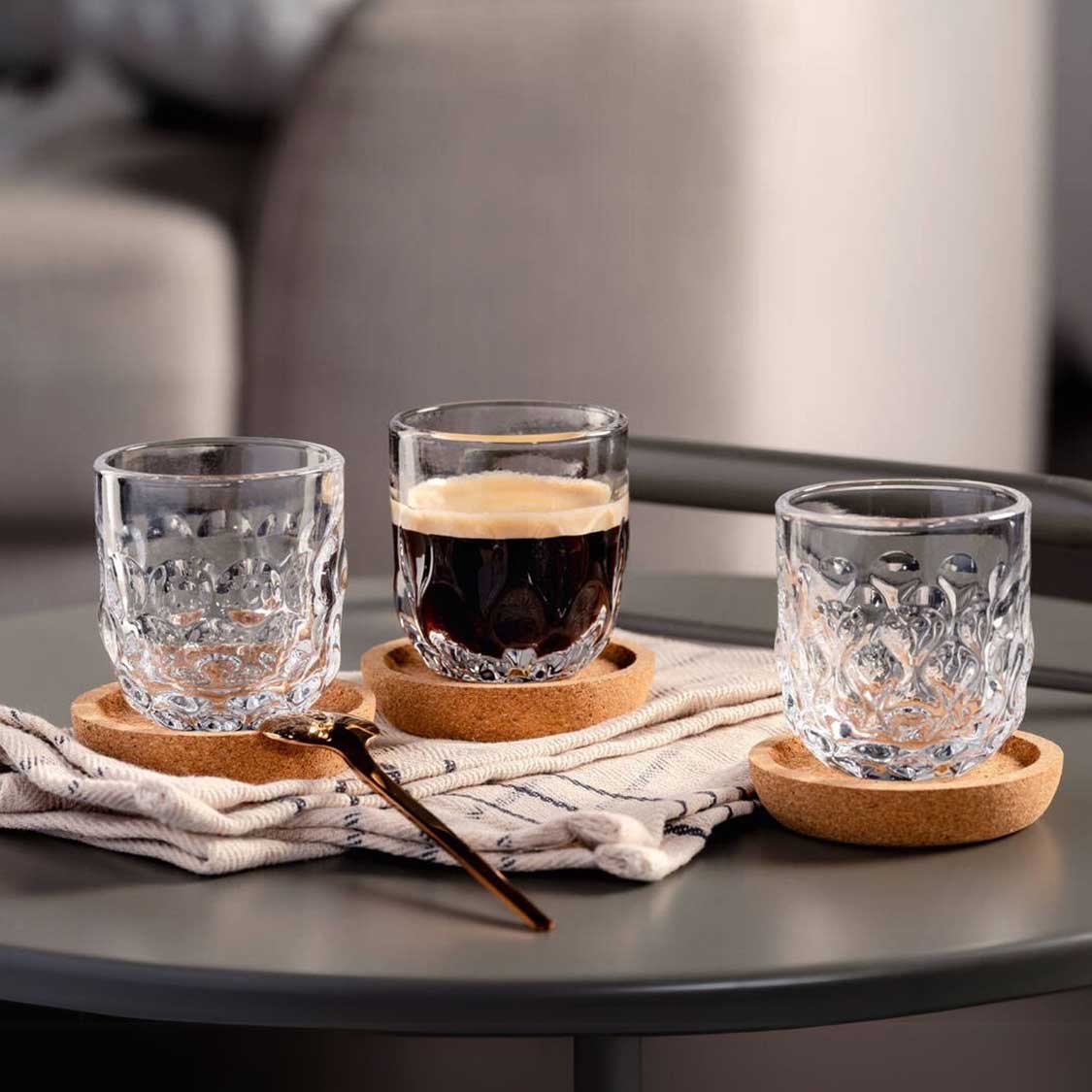 Espressoglas GOCCE auf Korkuntersetzer auf schwarzem Tisch mit Wasser und Kaffee