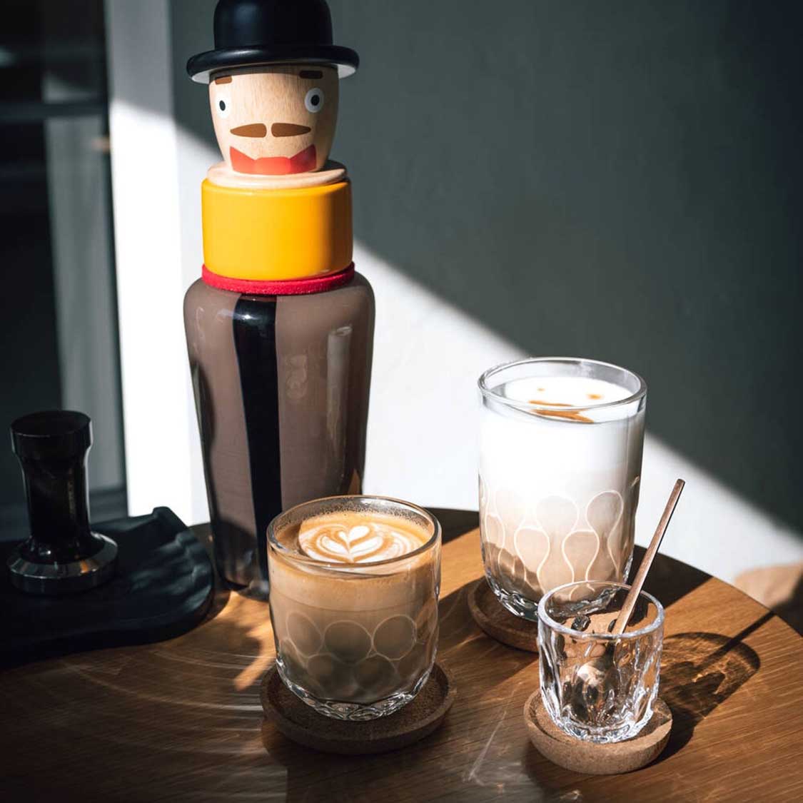 Espressoglas GOCCE mit Latte-Macchiato auf Holztisch, daneben Wasserglas und Dekofigur