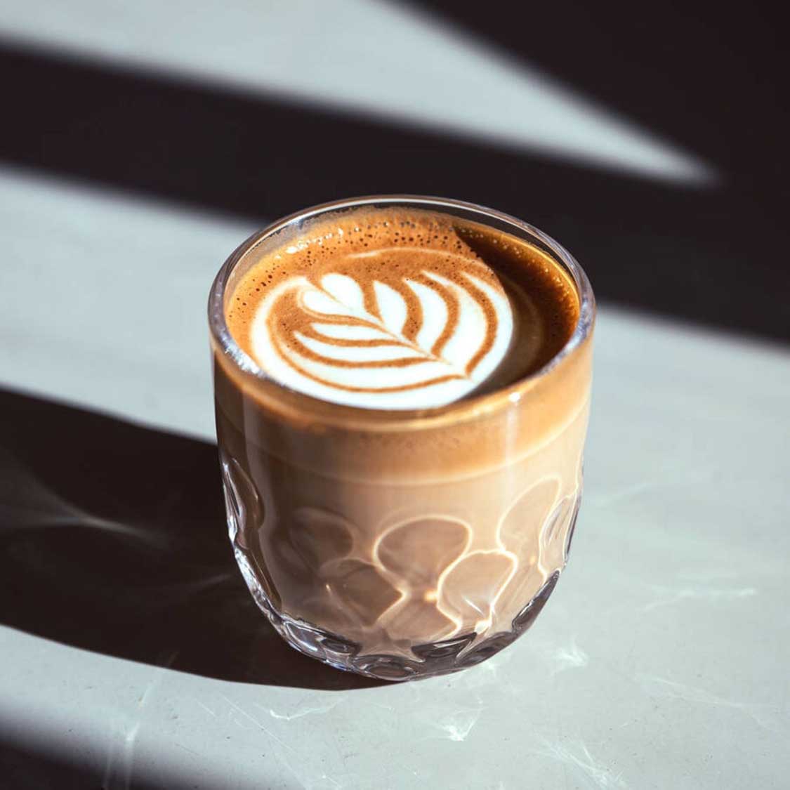 Kaffeeglas GOCCE mit Latte-Art-Motiv gefüllt, Sonnenlicht auf weißer Oberfläche