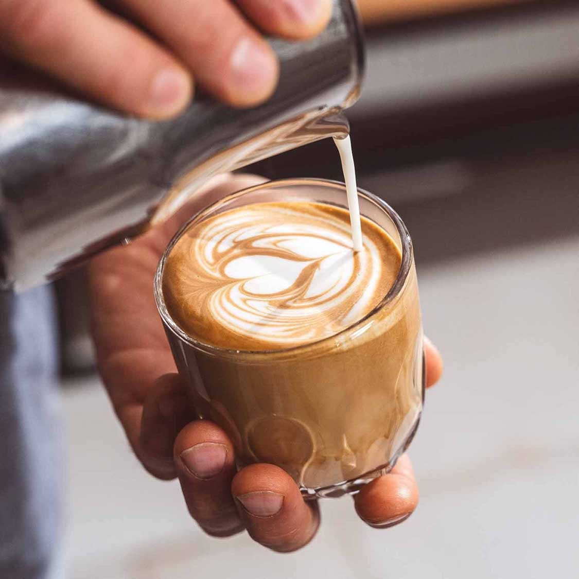 Kaffeeglas GOCCE 220ml mit Latte-Art, Milch wird eingegossen, Hand hält Glas