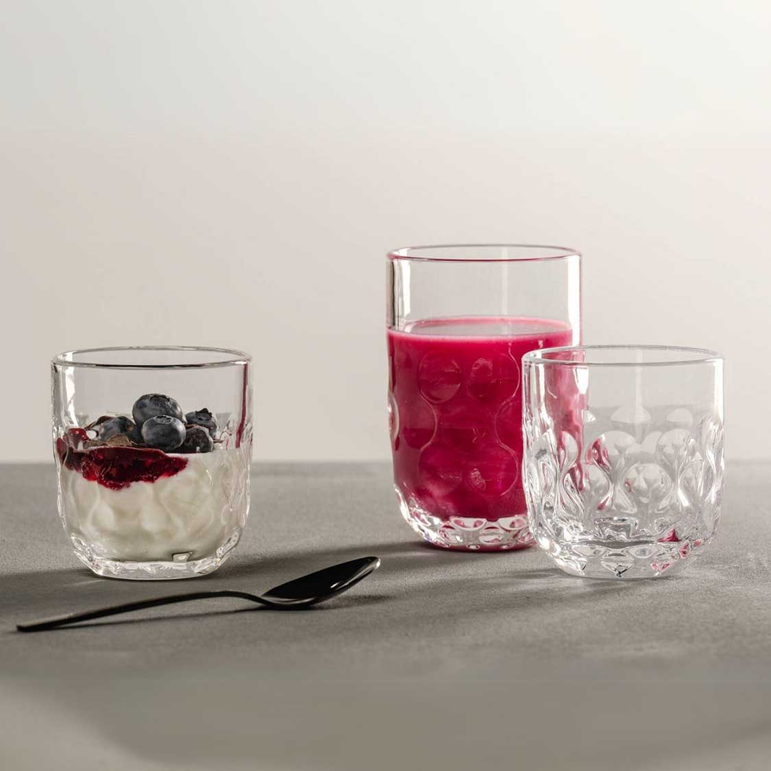 Latte Macchiato Gläser GOCCE mit Joghurt, Beeren und Saft auf grauer Oberfläche mit Löffel